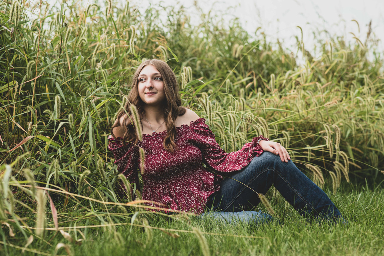senior girl in jeans and maroon top sitting among tall grass