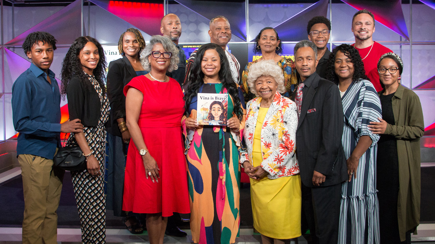 A group of diverse individuals posing together, one holding a book, all smiling in a studio setting.