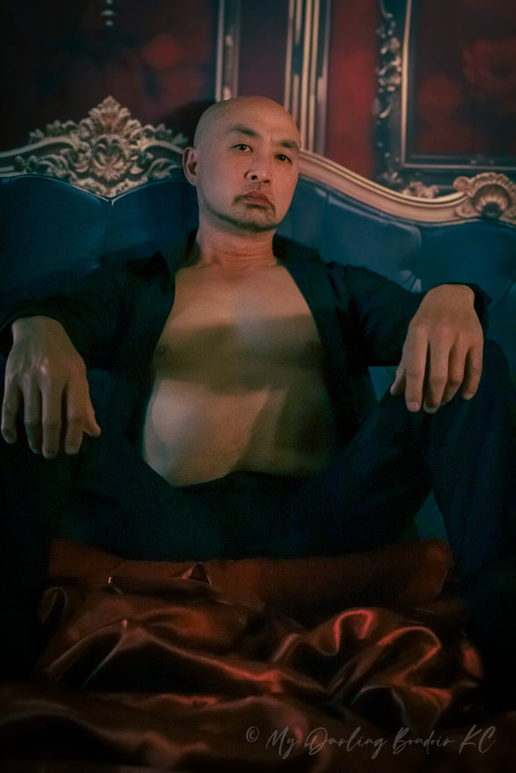 Man sitting on a luxurious blue couch in a dark, elegant room, wearing an open shirt.
