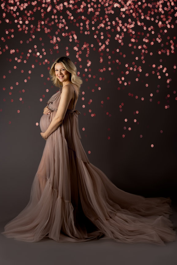 Pregnant woman in a flowing gown stands against a background of soft bokeh lights, showcasing her baby bump.