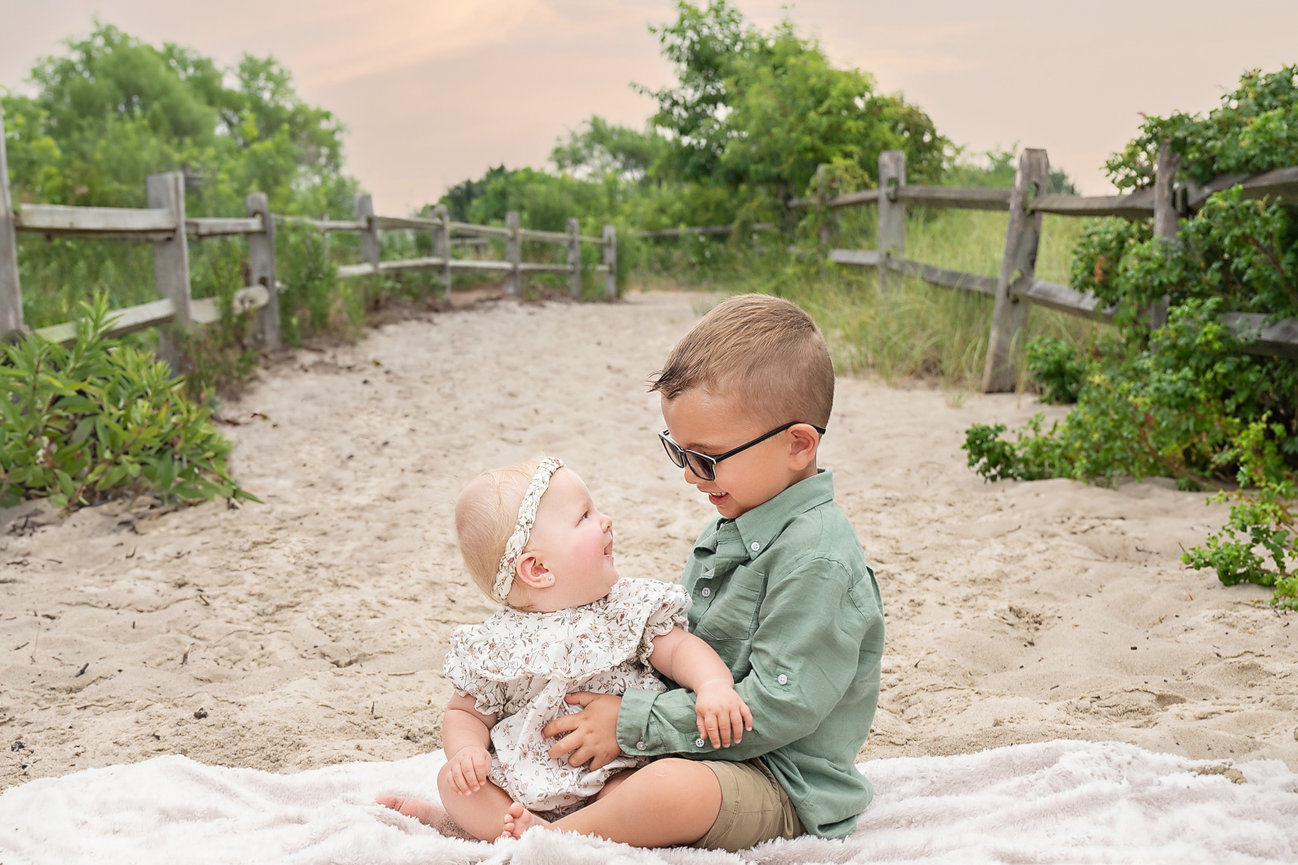 Toddler sitting in sand with big brother, laughing and simling