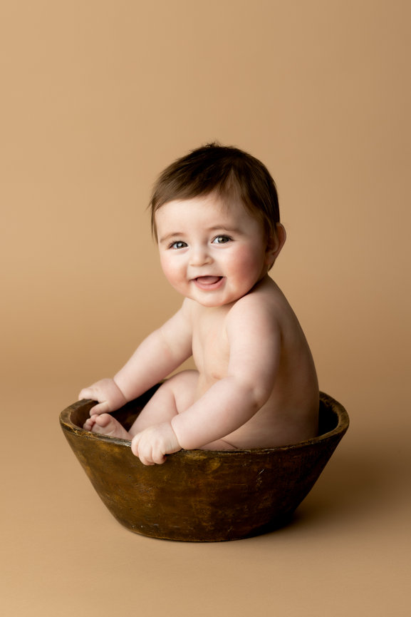 Baby sitting in a rustic bowl, smiling against a neutral beige background.