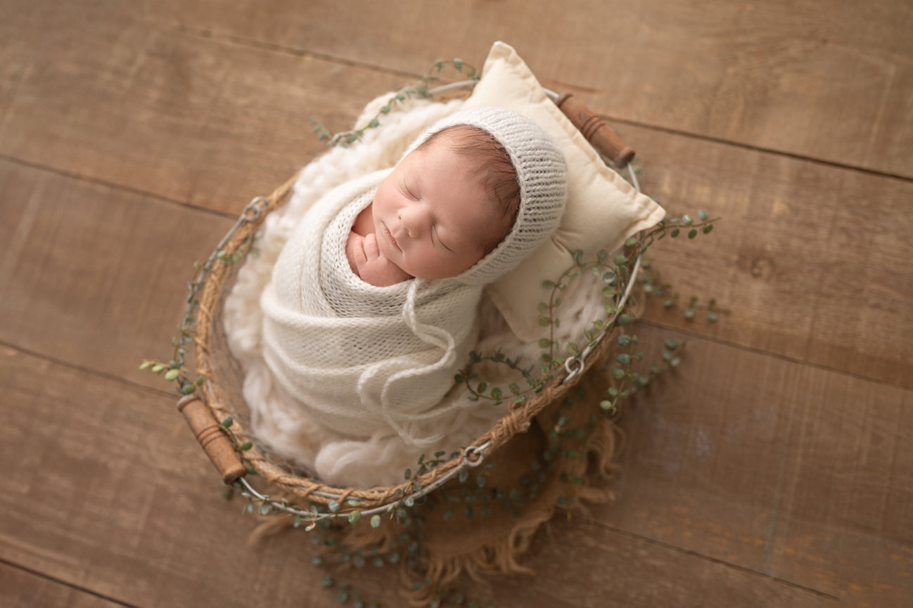 Cozy newborn portrait featuring baby wrapped in chunky knit swaddle and posed in shallow egg basket on white wood floor