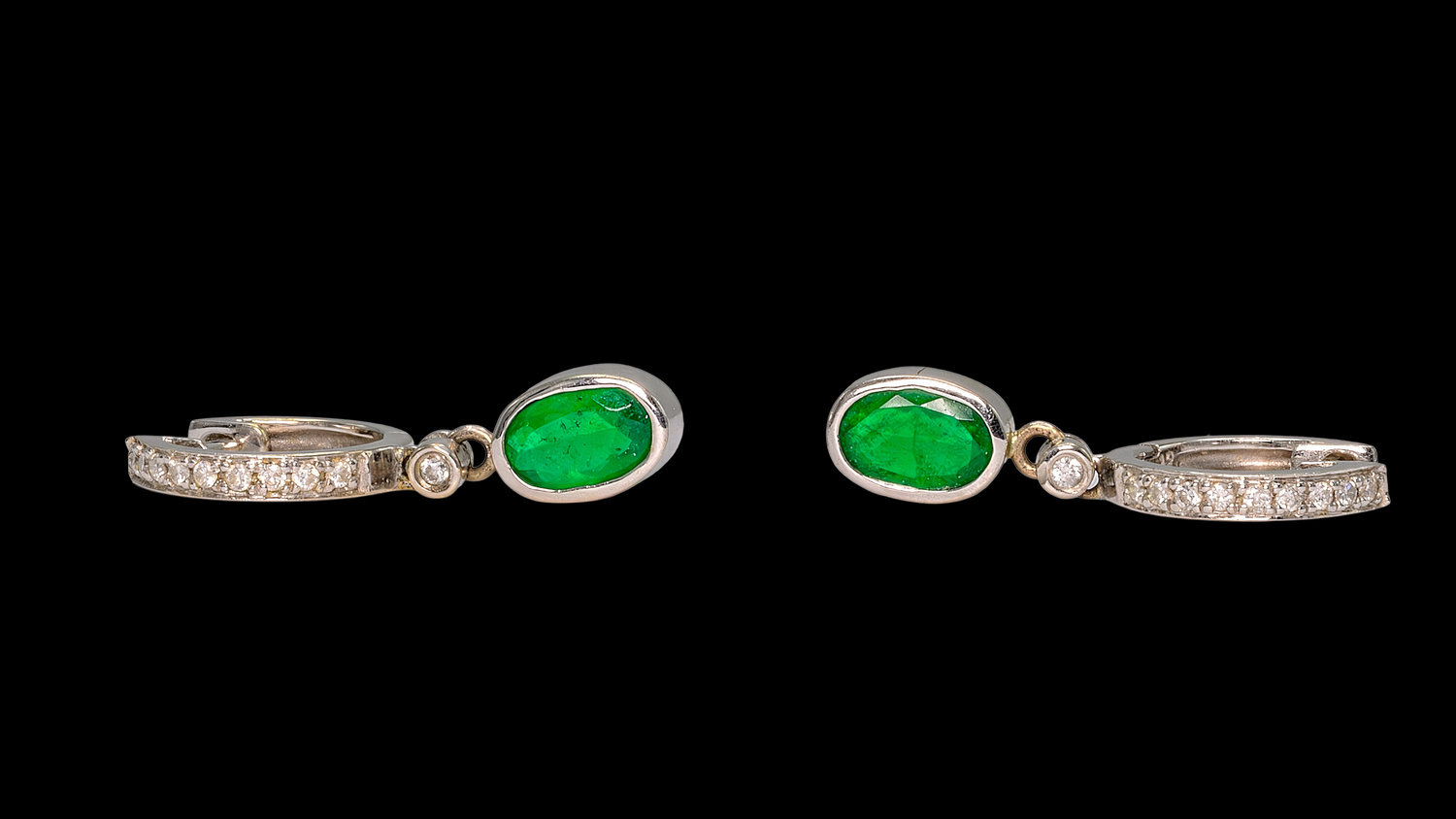 Pair of elegant drop earrings featuring oval-cut vivid green emeralds set in white metal bezels, accented by small round diamonds above the emeralds, with diamond-encrusted hoop settings, displayed against a solid black background