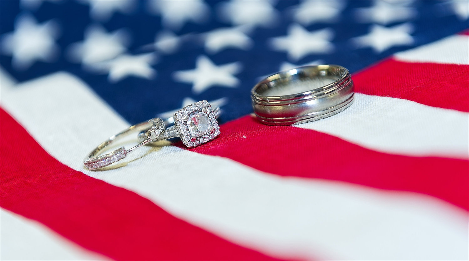 Diamond engagement ring with matching wedding band and a men’s wedding ring displayed on the fabric of the American flag.