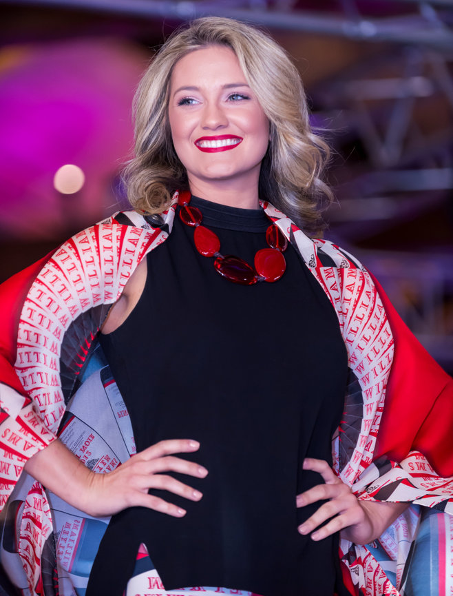 Smiling fashion model with blonde hair wearing a sleeveless black dress, bold red necklace, and a patterned red and white shawl, walking on a runway with a blurred purple and pink background.