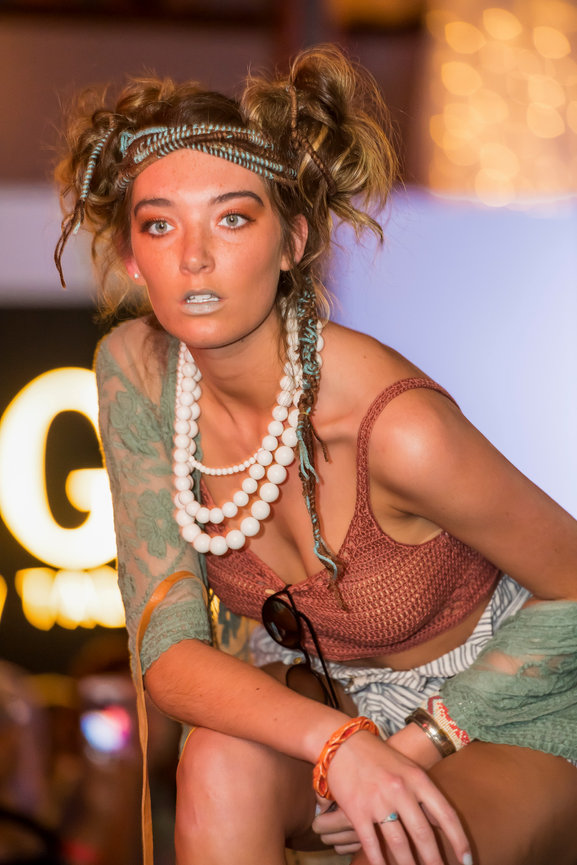 Runway model wearing a textured rust-colored crop top, striped shorts, and layered white bead necklaces with an eclectic hairstyle and turquoise braids, light metallic lipstick, and a sheer green floral shawl draped over her shoulders
