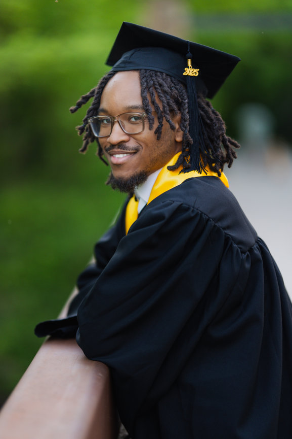 Young man in a graduation cap and gown, smiling confidently, outdoors with a greenery backdrop at Oak Point Nature Park.