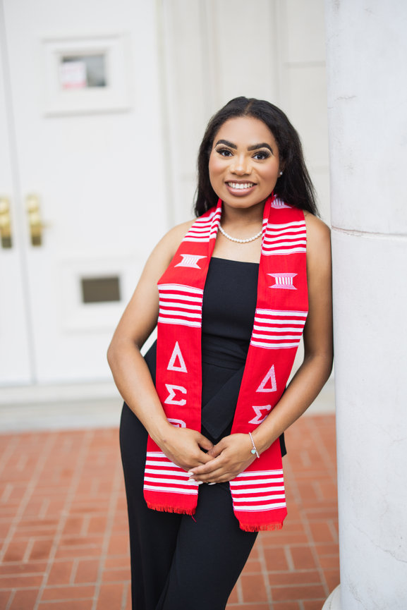 Graduate wearing a red and white graduation stole, smiling while leaning against a pillar at the University of North Texas