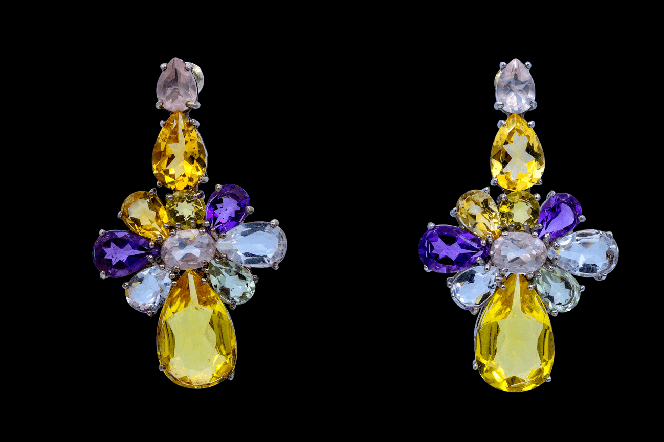 Pair of floral cluster drop earrings with multicolored gemstones. Features a pink oval center, surrounded by purple, yellow, green, and blue stones, finished with a large teardrop yellow gemstone, set in silver against a black background.
