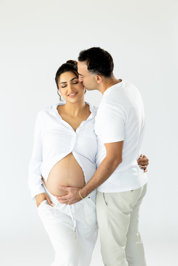 Pregnant woman in a white shirt, with a man kissing her forehead; both smiling and posing affectionately.