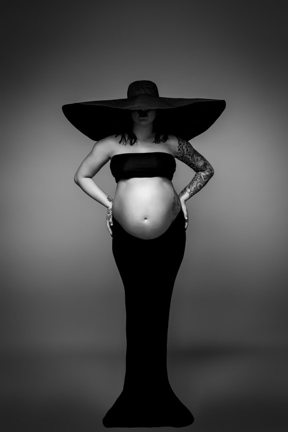 Pregnant person in a black bandeau and wide-brimmed hat, standing with hands on hips, monochrome.