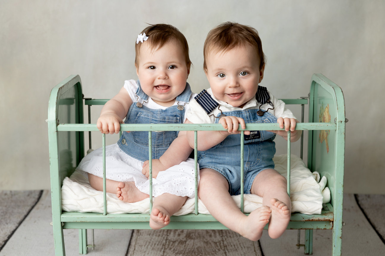 Two smiling babies in denim outfits sit together in a vintage green crib.