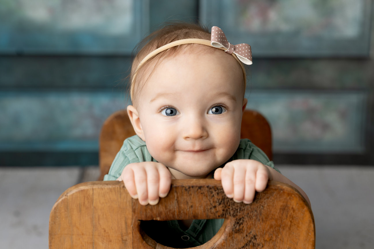 Smiling baby with a bow headband leaning on a wooden chair.