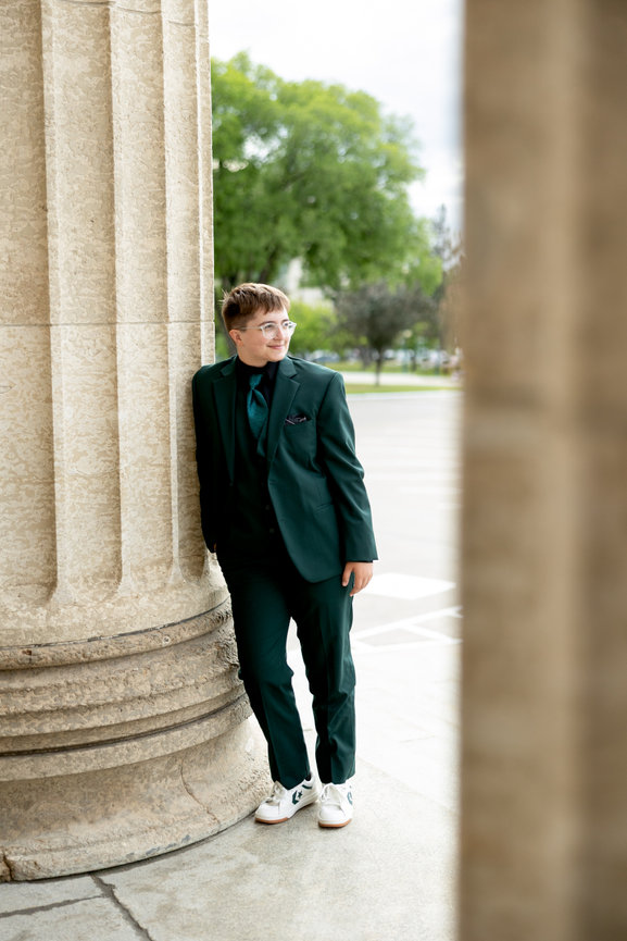 Person in a green suit leans against a stone column outdoors with trees in the background. Wearing glasses and white sneakers.