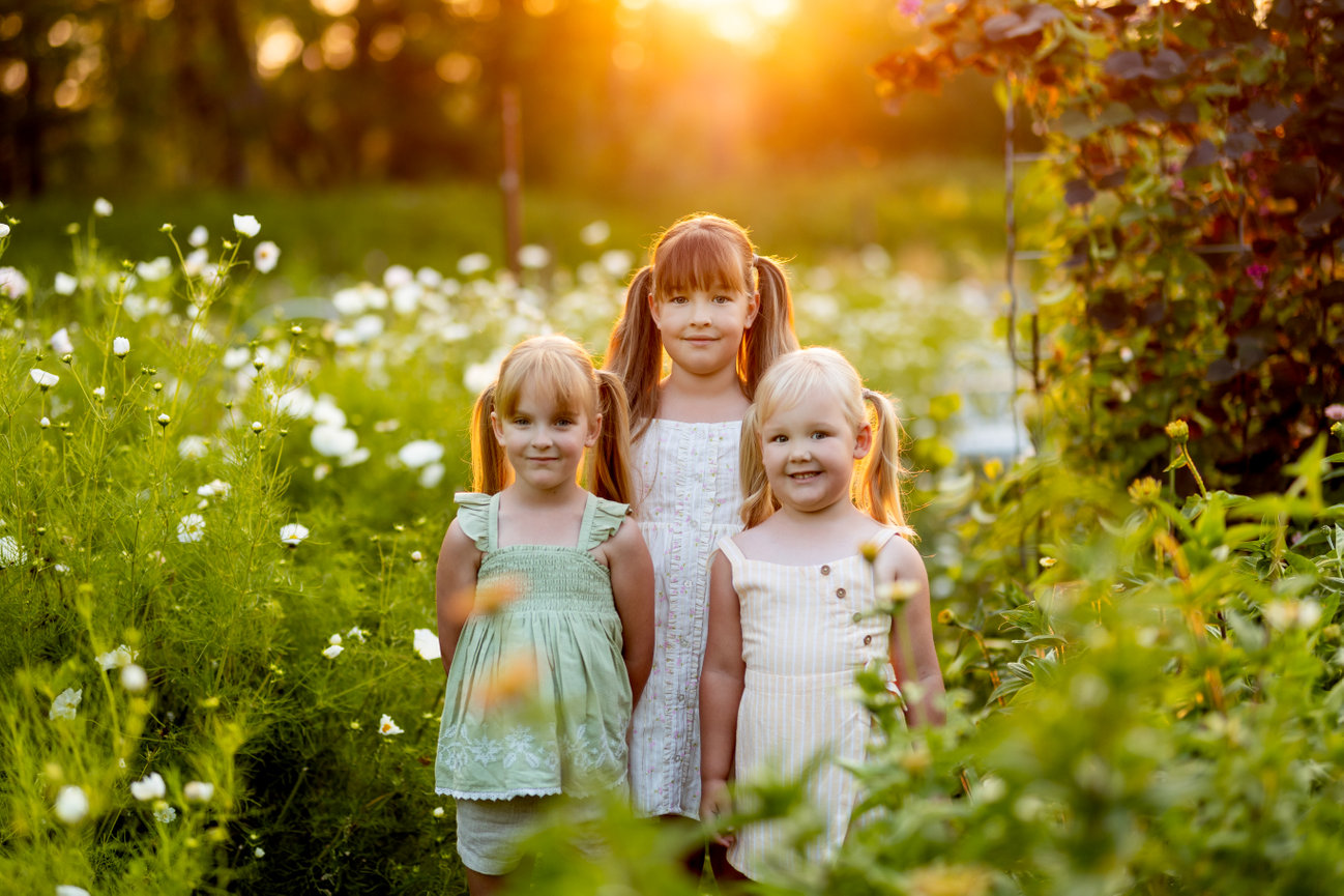 Three young children smiling in a sunlit garden with white flowers and greenery.
