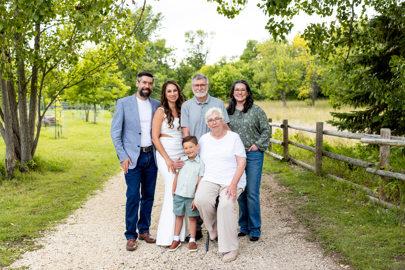 Family posing happily on a gravel path in a park, surrounded by green trees and a wooden fence.