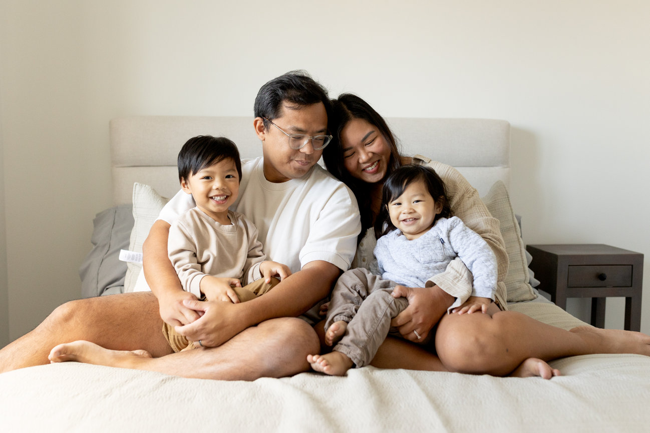 A family of four sitting on a bed, smiling warmly at the camera. Parents are in the middle, children on either side.