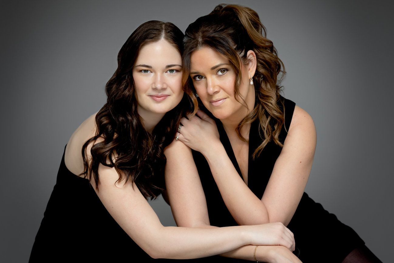 Two women with dark hair, dressed in black, pose closely together against a gray background.