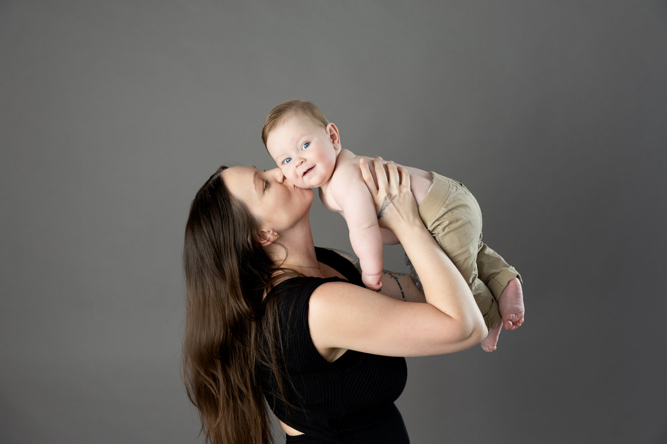 Woman kissing a smiling baby she's holding against a gray background.