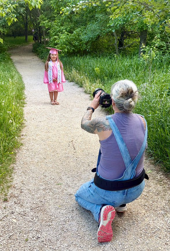 Girl in pink graduation cap and gown poses on a park path while a woman photographs her