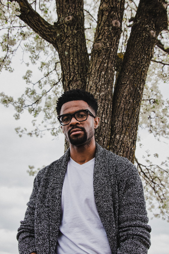 Man in glasses and sweater standing by a blossoming tree on a cloudy day.