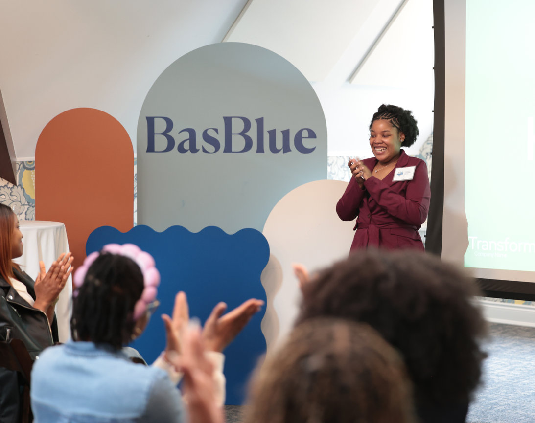 A woman speaks at a community event in front of a BasBlue sign.