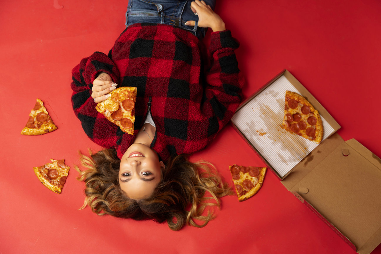 Fun senior portrait on red backdrop with teen eating pizza from open box.