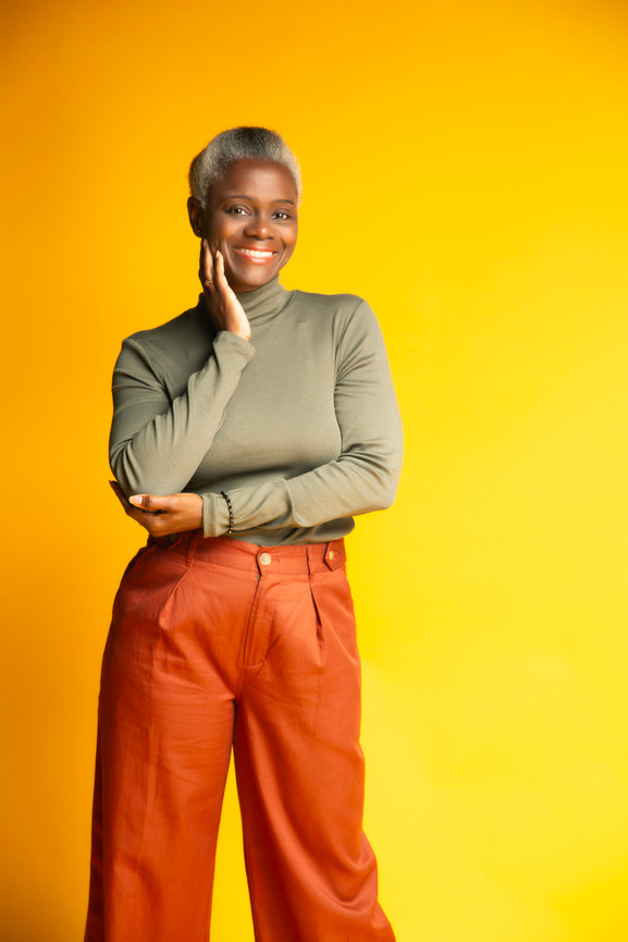 McKinney branding portrait of a stylish woman in orange pants and a green top, smiling brightly against a yellow backdrop.