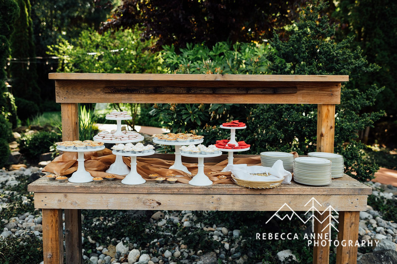 Outdoor dessert table with various pastries on white stands under wooden canopy, surrounded by greenery.