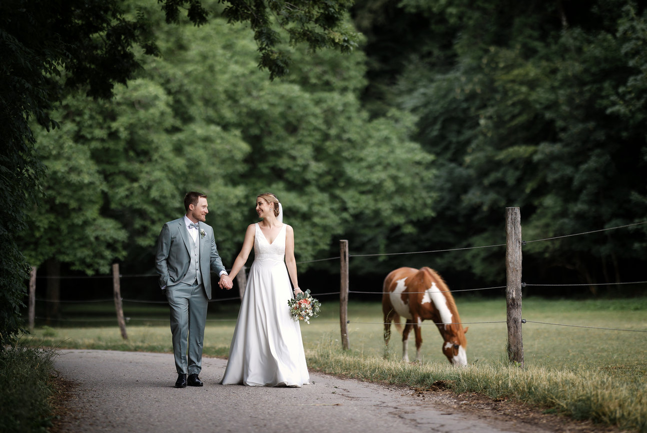 Bride and groom walking on a path beside a grazing horse in a wooded park