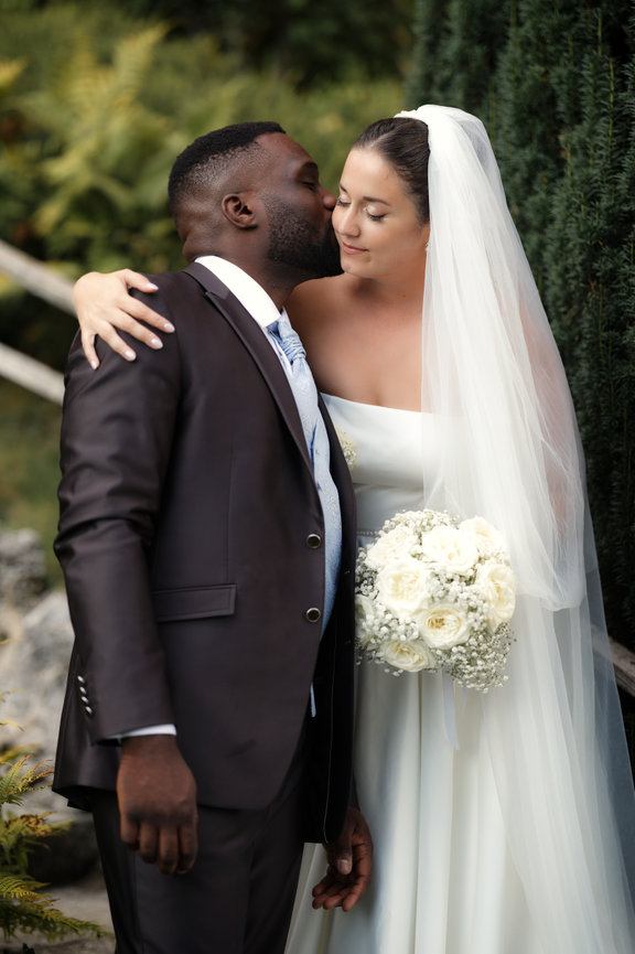 Wedding couple kissing, bride in white veil holding bouquet, groom in dark suit.
