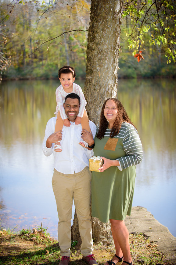 Family portrait by a lake, with a man holding a girl on his shoulders and a woman holding a gift.