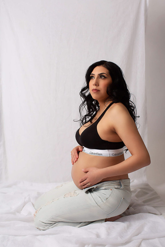 Pregnant woman sitting on knees on white fabric with white backdrop wearing jeans ad calvin klein bra photo taken during maternity photography session with kansas city maternity photographer Dreamworks Photography