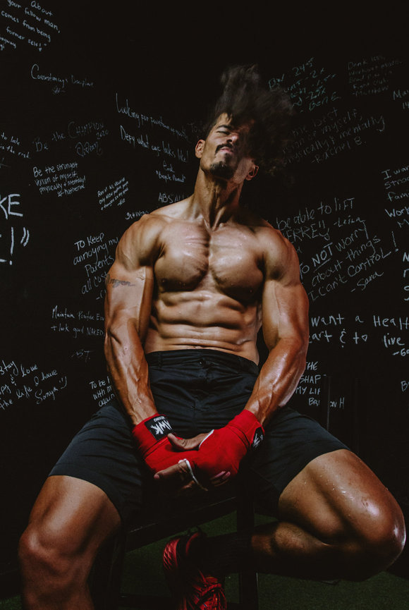 Athletic man with muscles sitting on stool flexing and grimacing with no shirt on and red gloves Female Athlete jumping and looking like she is running in mid air with a sport field in backdrop in photo by Sara Racey Photography