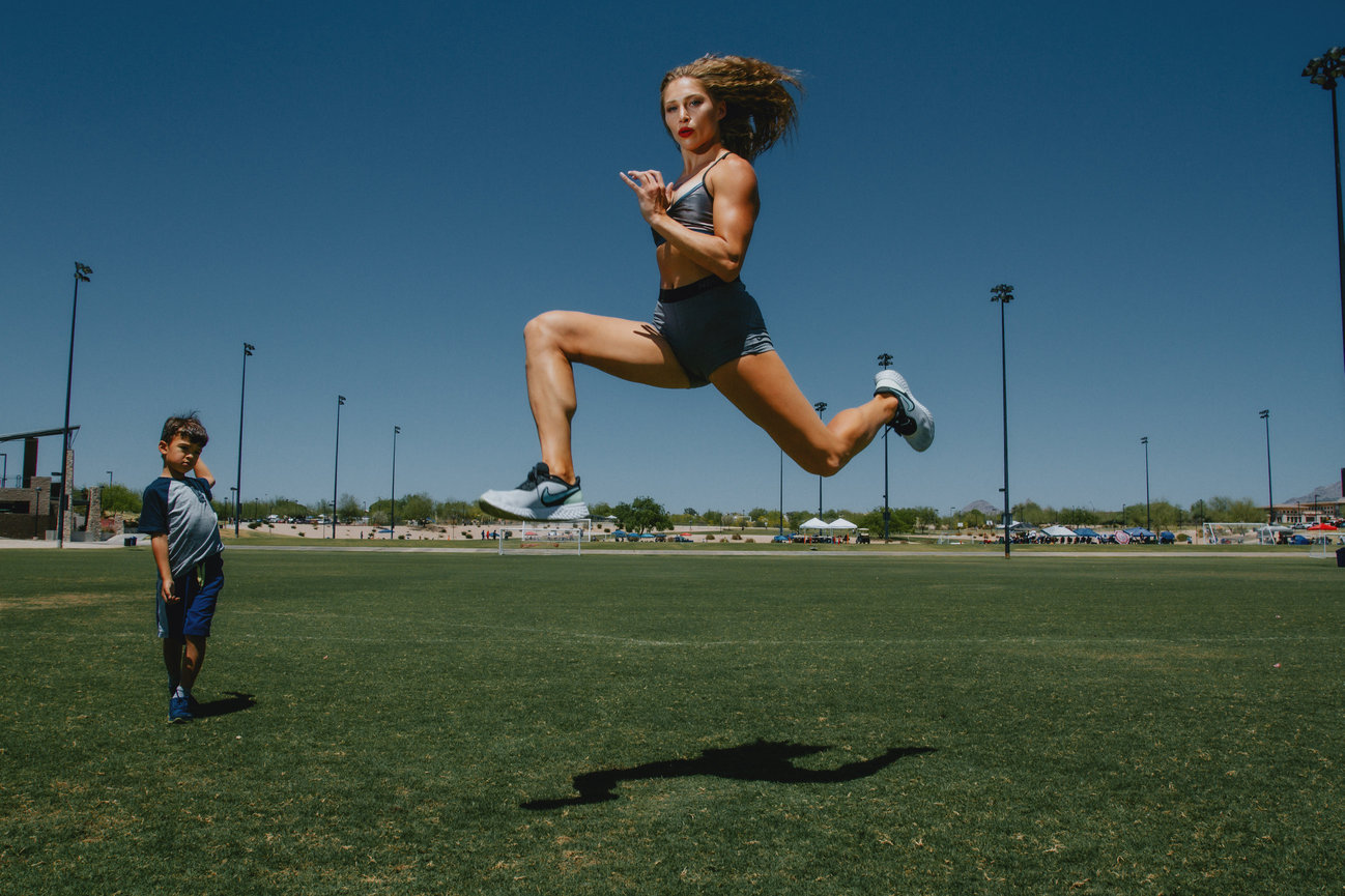 Female Athlete jumping and looking like she is running in mid air with the little boy watching with a sport field in backdrop in photo by Sara Racey Photographer.
