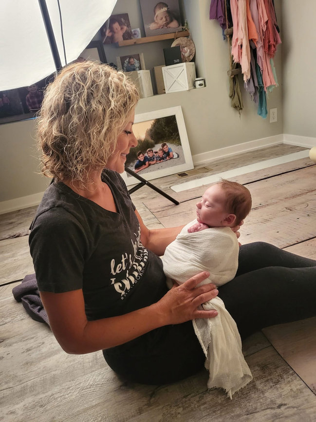Omaha Newborn Photographer holding a swaddled baby in a photography studio with family photos in the background.
