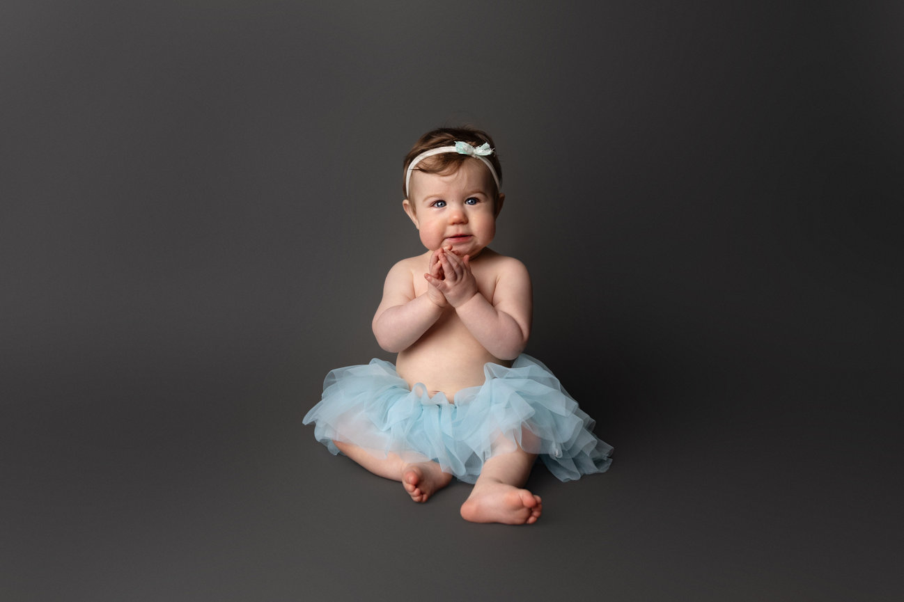 Baby in blue tutu and headband sitting against a gray background.