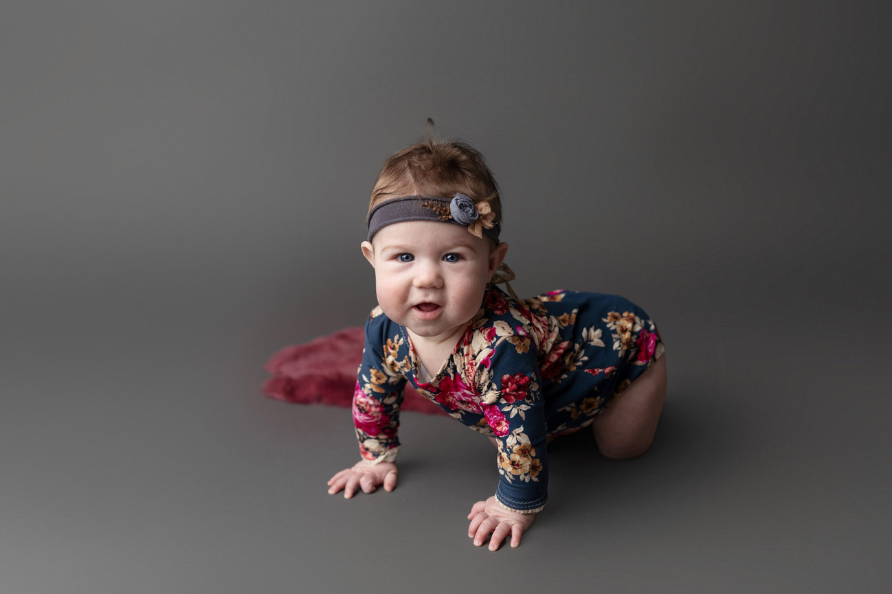 A baby in a floral outfit and headband is crawling on a gray background.