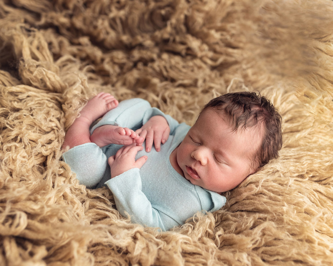 A newborn in a blue outfit sleeps peacefully on a fluffy beige blanket.
