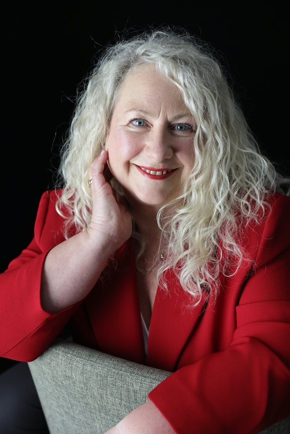 Smiling woman with curly blonde hair wearing a red blazer against a black background. authentic photos for my business and website