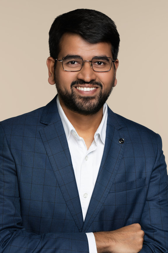 Smiling man in a blue blazer and glasses, standing with arms crossed against a neutral background.
