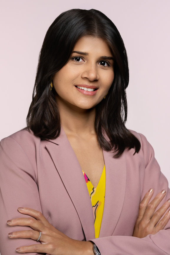 Professional headshot of a woman with long dark hair, wearing a pink blazer and yellow top, smiling confidently.