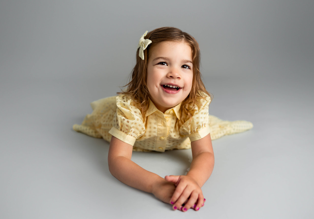 Toddler smiling in studio wearing bow and yellow, colorful outfit with soft background