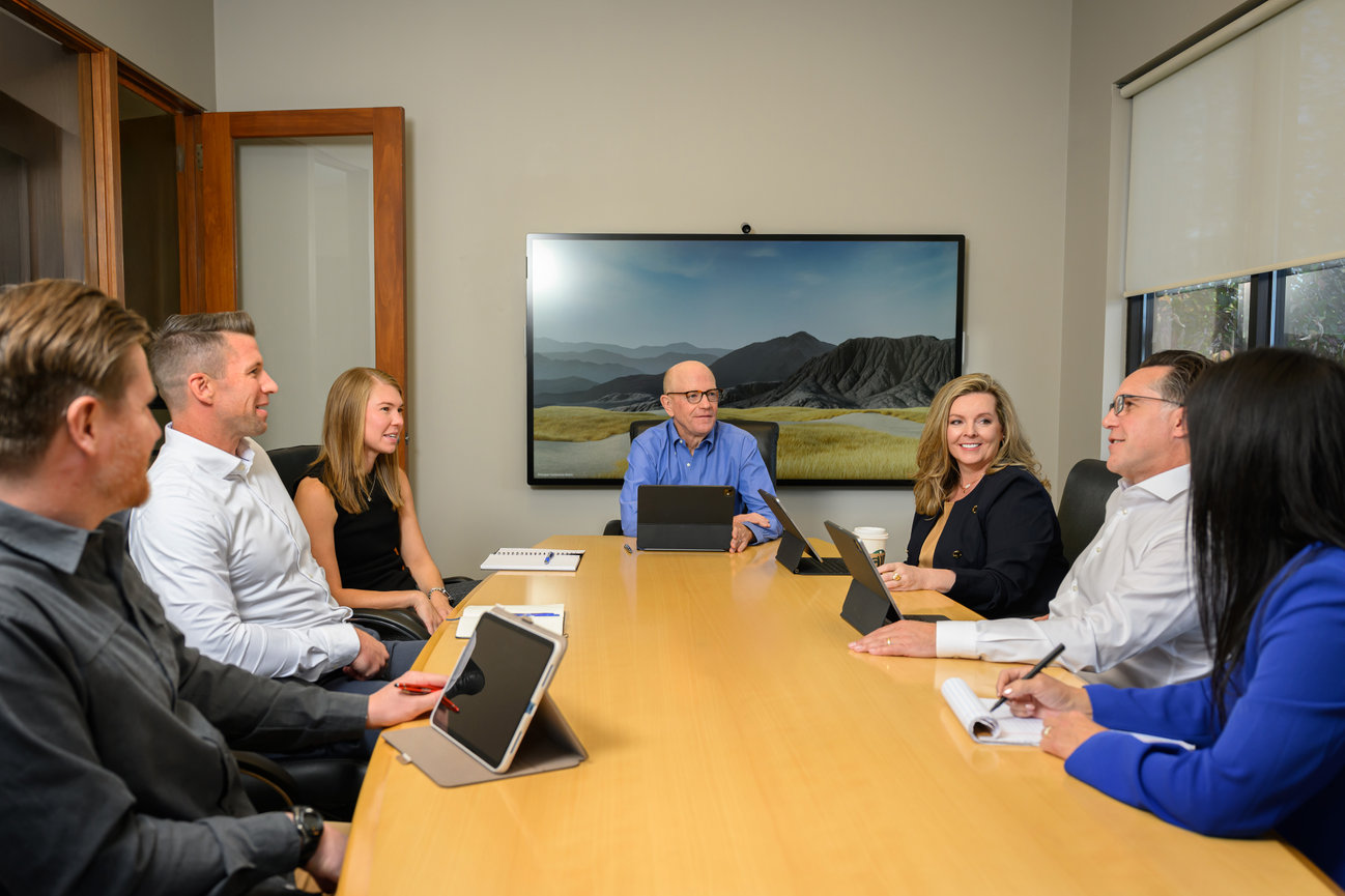 A diverse group of professionals in a conference room engaged in a meeting, with laptops and tablets on a table.