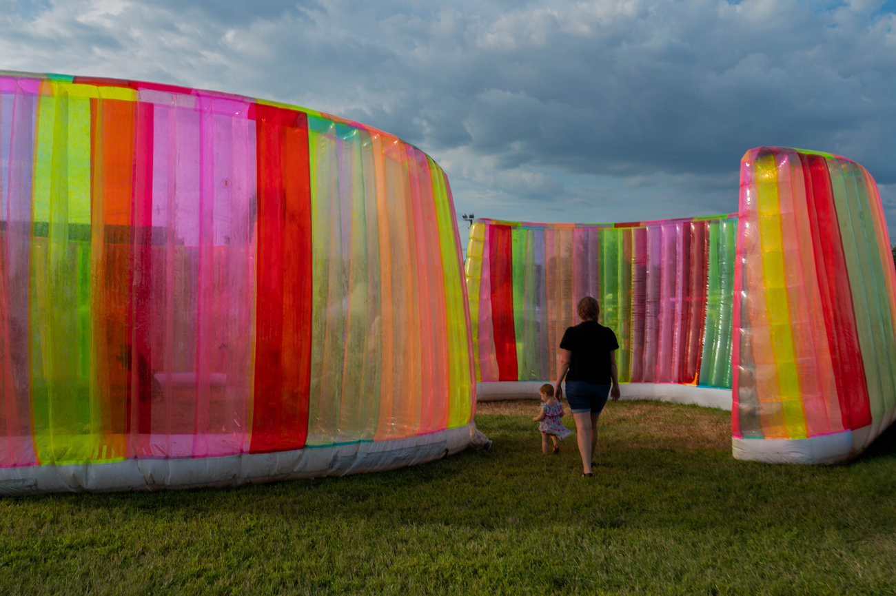 Woman and child walk through colorful, translucent inflatable structures in a grassy field under a cloudy sky captured in style of jo hayes images charlotte event photography