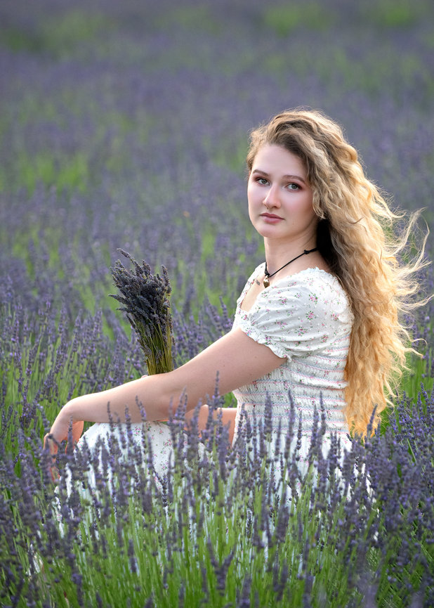 Portrait of a young blonde woman wearing a white dress, sitting in a lavender field holding a spray of lavender, and looking at the camera.