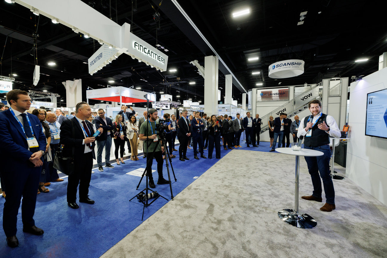 Speaker presenting to a crowd at a trade show booth with cameras and company signage