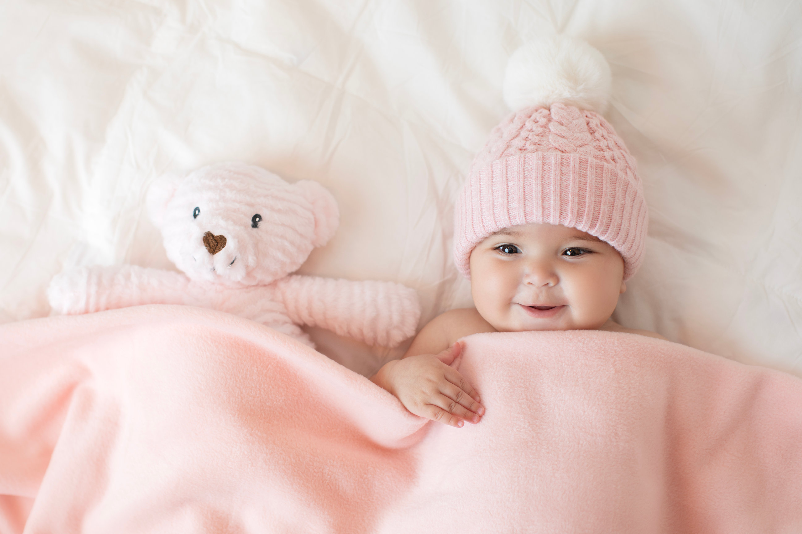 Baby in a pink hat peeking out from a pink blanket beside a teddy bear