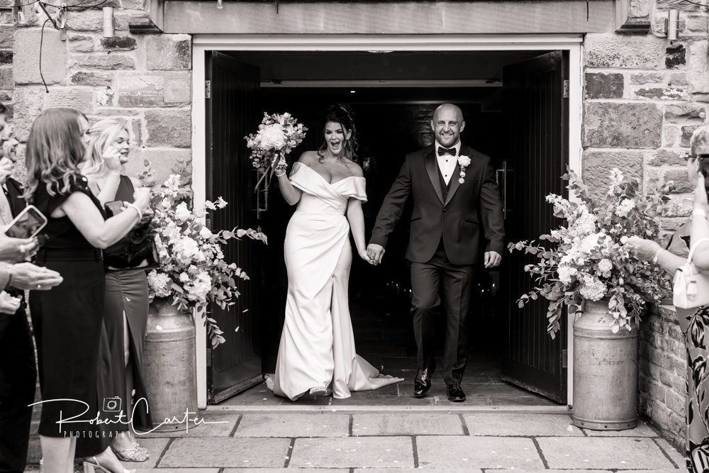 Bride and groom exit a stone building, holding hands, surrounded by applauding guests and floral arrangements.